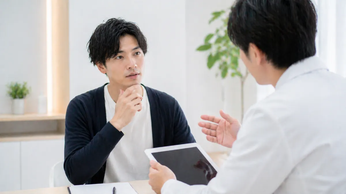 20代～30代日本人男性が少し不安そうな表情でクリニックのカウンセリングを受け、明るく清潔感のある空間でスタッフの説明を聞いている写真