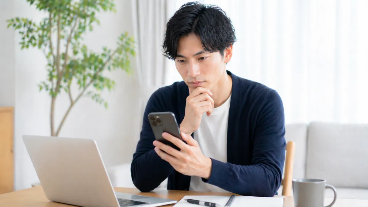 20代~30代日本人男性がスマートフォンで料金比較をしながら、テーブルの上のノートPCとメモ帳を前に真剣な表情でコスパを検討している清潔感ある室内の写真