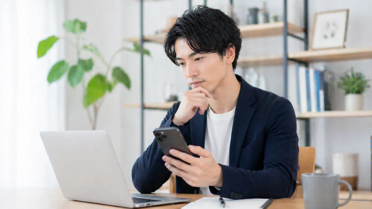 20代～30代日本人男性がスマートフォンとノートPCを使い、料金比較をしながら真剣な表情で検討している室内の写真