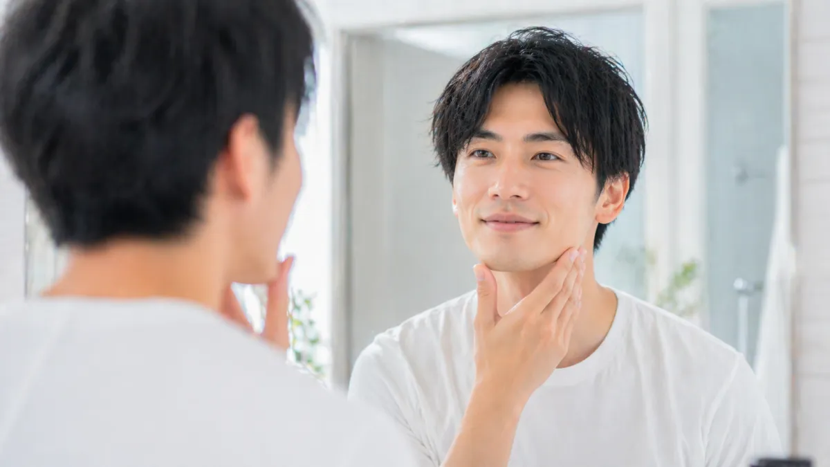 20代~30代日本人男性が明るく清潔感のある洗面所で鏡の前に立ち、ヒゲ剃り後のようなきれいな肌で爽やかに微笑みながら自信に満ちた表情を見せる写真